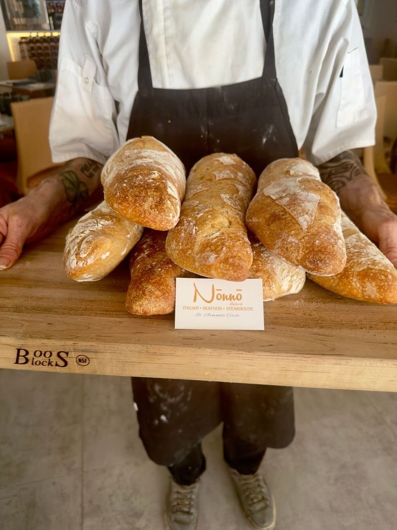 A baker holding a board of freshly baked bread with a Nōnnō card.