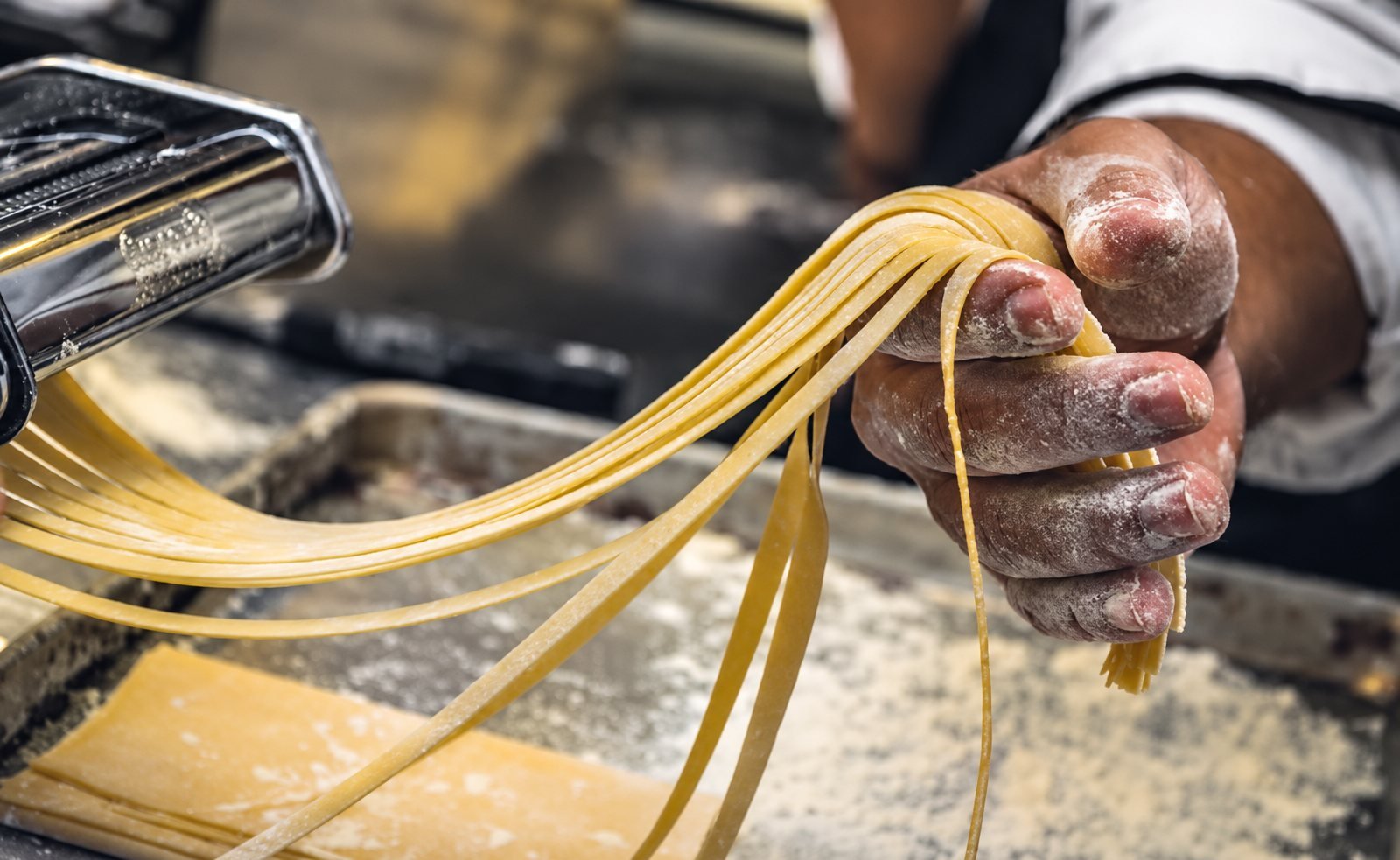 Flour-dusted hands lifting freshly cut pasta from a pasta machine.